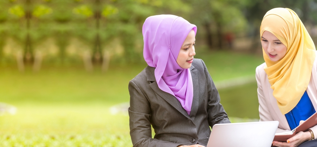 Photo of two women in office attire and hijabs talking together; one has an open laptop and the other a book