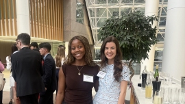 A Black woman and a white woman posing together and smiling for the camera at a networking event in an office reception room, with other attendees behind them and a drinks table to one side