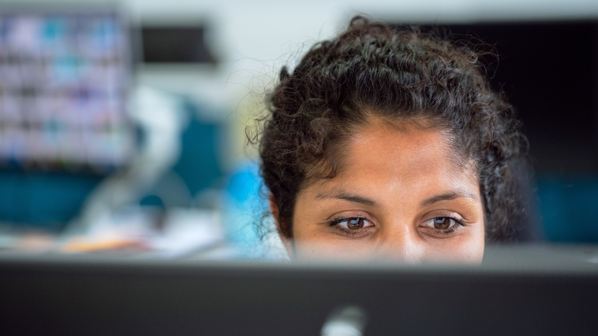 Young woman with dark curly hair working in front of a screen which is obscuring the lower part of her face