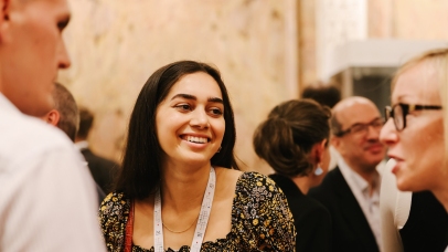 Photo of a woman in her 20s. She is at an event talking to two others, and there are more people in the background.