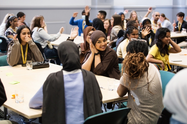 A groups of young people sat at tables, many smiling and with their hands raised.