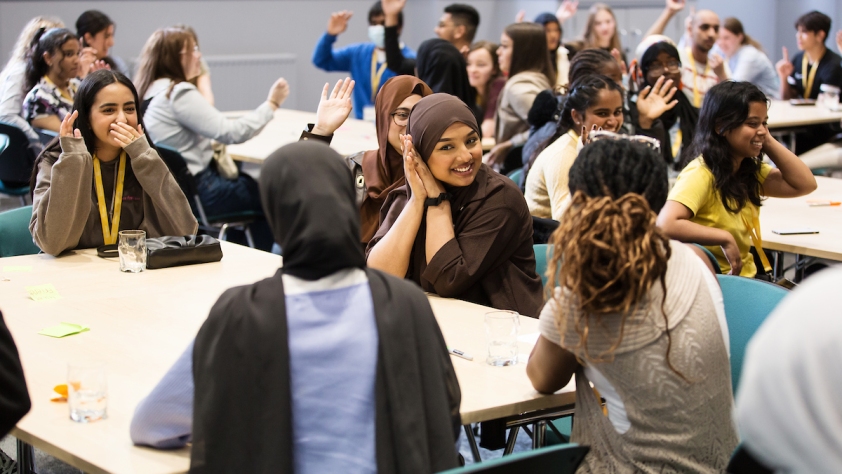 A groups of young people sat at tables, many smiling and with their hands raised.