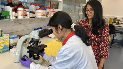 A young woman wearing a lab coat and sitting on a lab stool looking through a microscope, with a mentor standing beside her smiling.