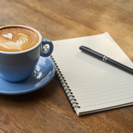 A cappuccino in a blue cup on a wooden surface next to a notebook and pen