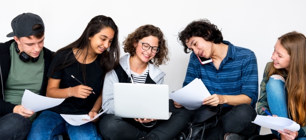 A group of students, of different genders and ethnicities, with papers and pens, a laptop and a phone, comparing notes together informally