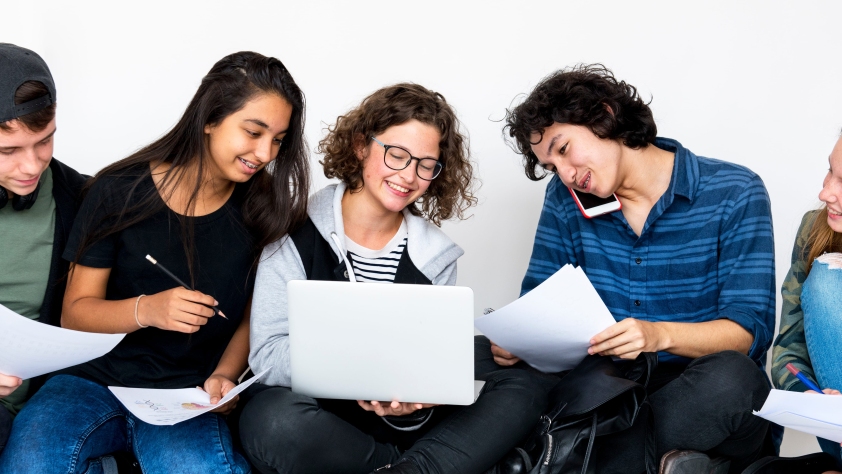 A group of students, of different genders and ethnicities, with papers and pens, a laptop and a phone, comparing notes together informally