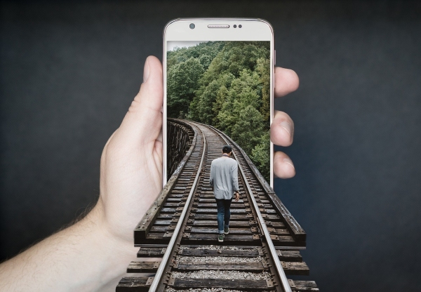 Photo of a hand holding out a smartphone. A railway track extends from an image on the screen out towards us and there is a man walking down the track into the screen.