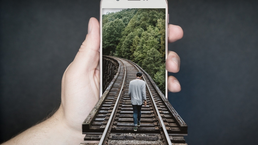 Photo of a hand holding out a smartphone. A railway track extends from an image on the screen out towards us and there is a man walking down the track into the screen.