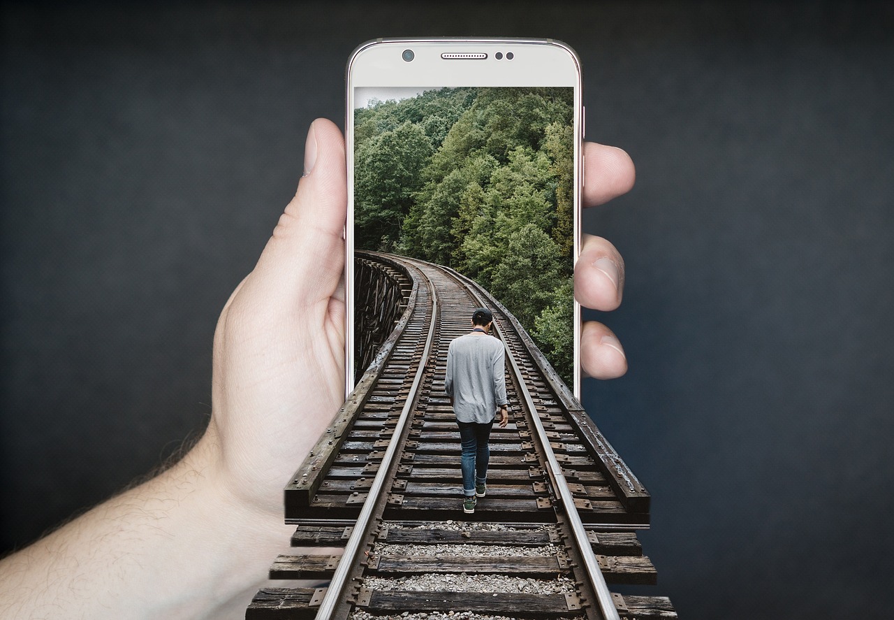 Photo of a hand holding out a smartphone. A railway track extends from an image on the screen out towards us and there is a man walking down the track into the screen.